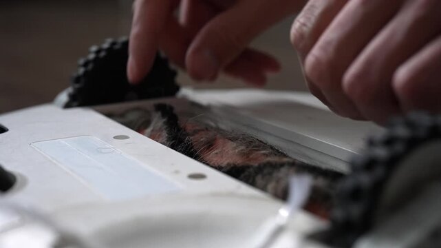 Close-up of person hand removing hair, dust, debris from robotic vacuum cleaner roller brush. Man performs maintenance home appliance, cleaning parts for better performance. Shooting in slow motion.