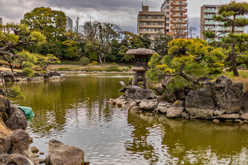 Fototapeta premium Grey heron by the stone lantern contrasting with urban buildings at Kiyosumi Gardens March 2024