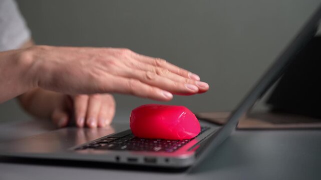 Close-up of hand pressing pink cleaning gel onto laptop keyboard. Person using sticky slime putty to remove dust and dirt from between computer keys. Modern gadget hygiene and maintenance concept.
