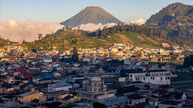 Aerial view of the town of Kejajar nestled in the Dieng plateau with terraced hillsides, a prominent mosque, and a volcanic mountain peak in the background in Kejajar, Central Java, Indonesia.