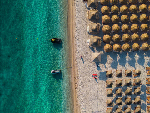 Aerial view of Llamani Beach with rows of straw umbrellas on the sandy shore and jet skis floating in the turquoise Ionian Sea in Himare, Vlore County, Albania.