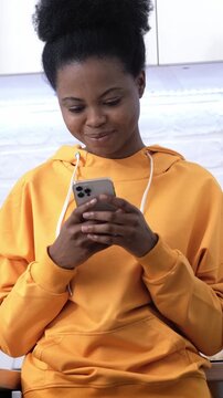 Cheerful young African American woman holding smartphone celebrating success, freelance work and online study concept, happy student at home.