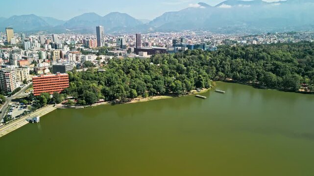 Albania, Tirana 06 09 2025: Arc drone video orbiting Grand Park lake with trees, city center skyline, Arena Kombetare stadium, Marriott hotel tower and mountains on sunny day.