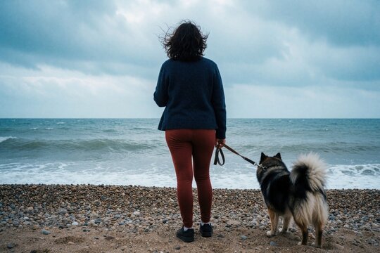 Woman with her dog on a pebble beach looks out at the wavy sea under a cloudy sky. They stand on the shore, enjoying a quiet moment together.
