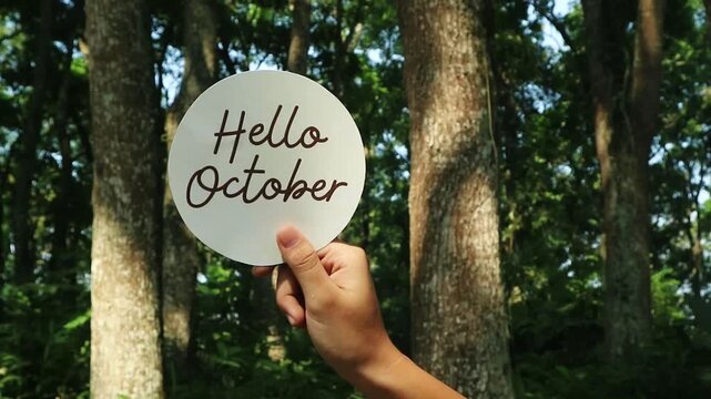 A hand holding a circular sign with the text Hello October in a forest setting.