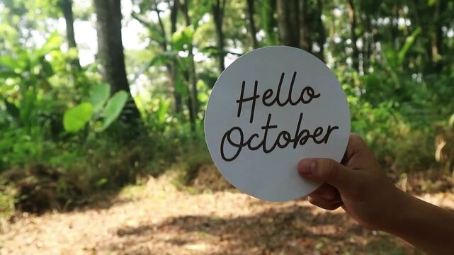A person holding a circular sign that says Hello October in a lush forest.