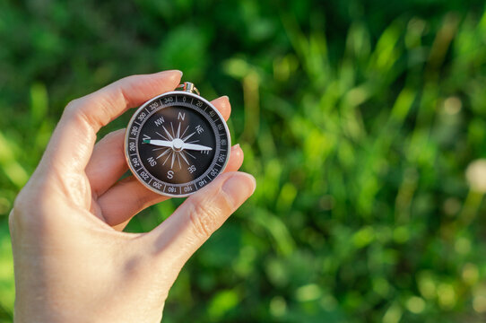 Hand holding a magnetic compass against a blurred green background, symbolizing navigation, direction, travel, adventure and finding the right path in nature.