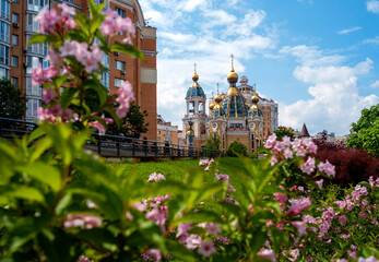 Scenic view of the Nativity of Christ Church on the Obolon waterfront in Kyiv, Ukraine, with distinctive golden domes framed by blooming pink flowers and modern urban architecture. © maxsyd
