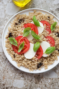 Caprese salad served with quinoa on a white plate, vertical shot on a light-brown granite background, middle closeup, elevated view