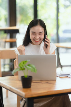 A cheerful young Asian businesswoman celebrating success while talking on the smartphone and working on a laptop at the desk in a modern office.