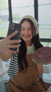 Happy female potter taking selfie with smartphone while holding handmade ceramic bowl in pottery studio, creative artisan and small business lifestyle