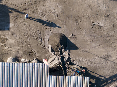 Aerial view of a stone crushing operation with a conveyor belt, a pile of crushed stone, and a worker walking across the dusty ground in Kecamatan Karangnongko, Jawa Tengah, Indonesia.