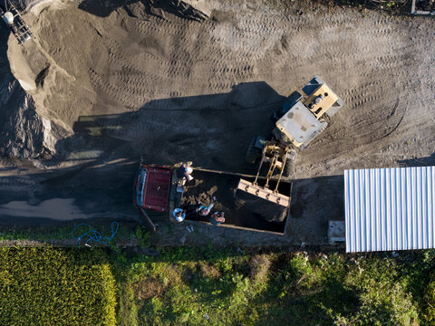 Aerial view of stone crushing site with yellow loader filling red truck, workers nearby, piles of gravel, and surrounding vegetation in Kecamatan Karangnongko, Jawa Tengah, Indonesia.