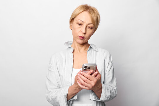 Woman holding smartphone in studio, looking down with thoughtful expression, lips slightly pursed, white background, soft light, modern technology communication