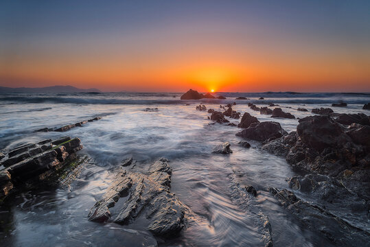 Vibrant sunset over the rocky coast of Barrika beach with long exposure sea water flowing between flysch rocks and golden glowing sun