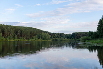 Obraz premium Calm river flows through a lush forest, reflecting trees and clouds under a clear blue sky, creating a tranquil natural scene