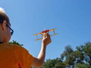 A boy plays airplane pilot with a wooden toy airplane, holding it in his hand against a blue sky background. © Prrrettty