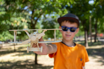A boy holds a wooden toy airplane with a propeller on the front at arm's length while sitting outside in a park. © Prrrettty