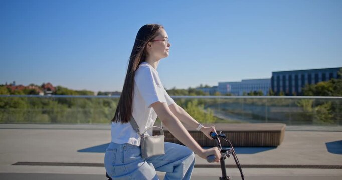 A young beautiful girl in casual clothes rides a bicycle on a bridge in the city in the morning during blue hour