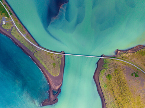 Aerial view of a bridge spanning a turquoise river with dark volcanic sand banks and green terrain in Iceland.