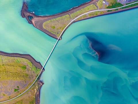 Aerial view of a bridge spanning a turquoise glacial river with black sand banks and green coastal vegetation in Iceland.