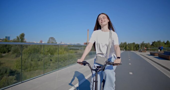 A young beautiful girl in casual clothes rides a bicycle on a bridge in the city in the morning during blue hour