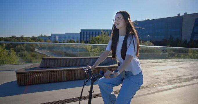 A young beautiful girl in casual clothes rides a bicycle on a bridge in the city in the morning during blue hour