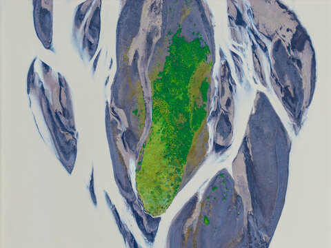 Aerial view of a vibrant green island surrounded by dark sediment and glacial meltwater in a braided river system in Iceland.