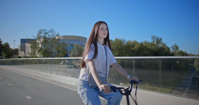 A young beautiful girl in casual clothes rides a bicycle on a bridge in the city in the morning during blue hour