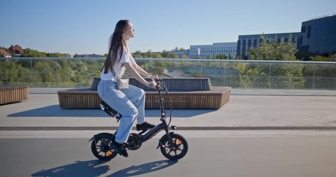 A young beautiful girl in casual clothes rides a bicycle on a bridge in the city in the morning during blue hour