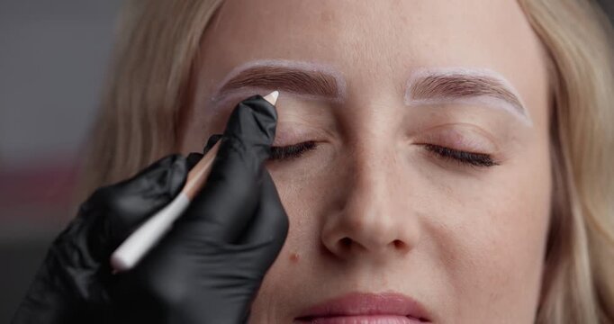 Handheld closeup of cosmetician drawing and mapping eyebrow stencil on woman face in beauty salon before permanent makeup.