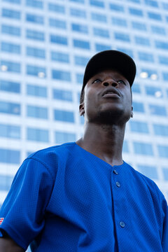 Young african american man in baseball jersey and cap looking up with determined expression against modern city building
