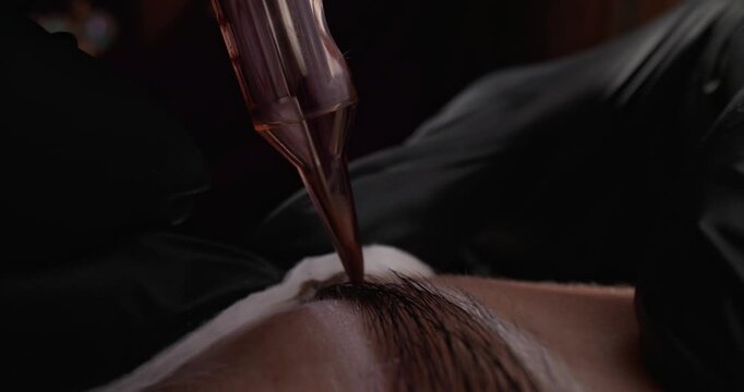Closeup microblading technician applying pigment to eyebrow on client's face in treatment studio.