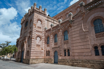 Exterior view of the Gran Teatro Falla, a historic red-brick theater in Cadiz, Spain, featuring neo-Mudéjar architectural style under a partly cloudy sky. © ihervas