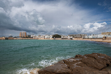 Wide coastal view of La Caleta Beach in Cádiz, Spain, with turquoise waters, rocky foreground and dramatic clouds over the historic shoreline and seaside buildings. © ihervas