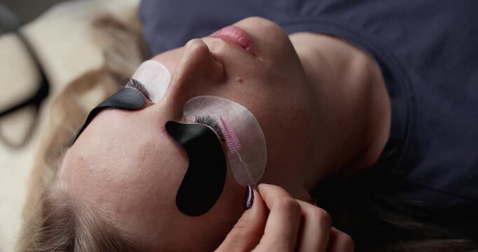Handheld closeup eyelash extension application on woman lying on treatment bed in salon with technician hand movement tracking.