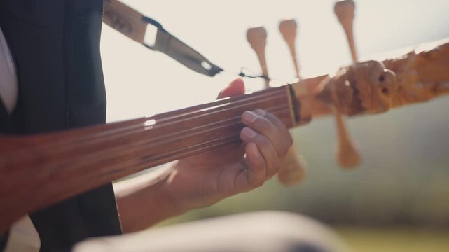 Close-up macro shot of a musician's hands tuning carved wooden pegs on a traditional Himalayan lute under warm outdoor sunlight