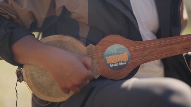 Nepali folk musician's hands plucking strings on a wooden lute with a leather-covered soundboard while seated outdoors in soft daylight