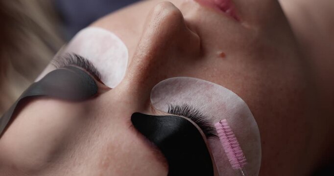 Closeup tracking shot of eyelash technician brushing and applying extensions on client's closed eyes during beauty salon treatment handheld gentle motion.
