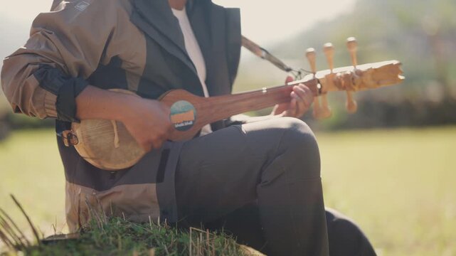 Wide shot of a traditional musician sitting on green grass playing a handmade lute in golden backlit sunlight in the Himalayan countryside
