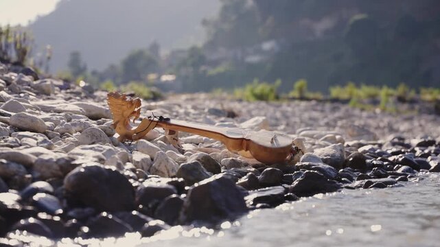 Close-up of a carved wooden lute placed at the edge of a flowing Himalayan river with sunlit water ripples and rocky terrain