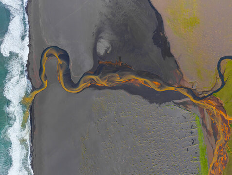 Aerial view of a winding river with golden sediment flowing across a vast black sand beach into the Atlantic Ocean in Iceland.