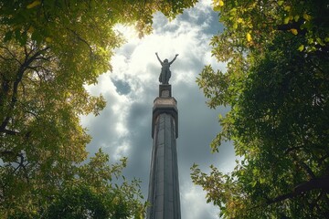 Obraz premium Tall stone column topped by a triumphant statue with raised arms framed by green tree canopy beneath dramatic sunlit clouds