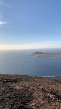 Panoramic view of La Graciosa Island from a high viewpoint in Lanzarote, Canary Islands, Spain. The landscape shows the volcanic island surrounded by the deep blue waters of the Atlantic Ocean, with i