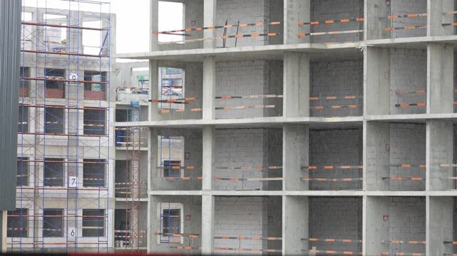 Concrete building frames form an incomplete structure. Scaffolding lines the left side of the construction site. Other buildings appear in the background. This footage shows early construction phases