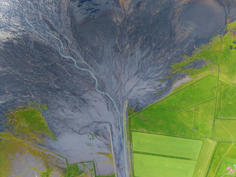 Aerial view of a glacial river delta with intricate dark volcanic sediment patterns flowing through vibrant green fields in Iceland.