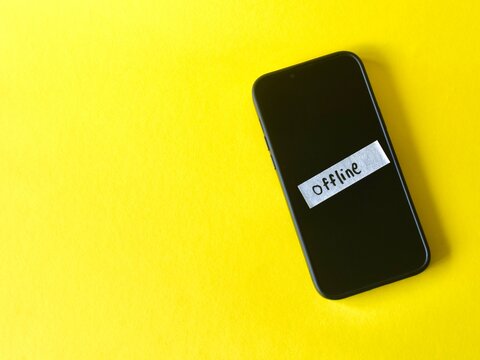 Flat lay of a smartphone on a yellow surface with a white strip of paper taped to the screen showing the handwritten word Offline.
