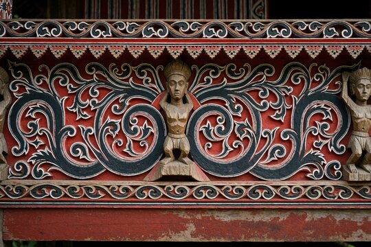 Traditional Batak Toba house with detailed Gorga wood carving. Ethnic Indonesian architecture features red black, white patterns. Ancient tribal ornament shows human figures, floral motifs on wooden