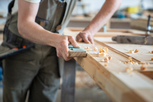Craftsman holding block with sandpaper and polishing surface of vintage wooden door at worktable, skillful man carpenter removing defects and preparing door for renovation in restoration workshop.