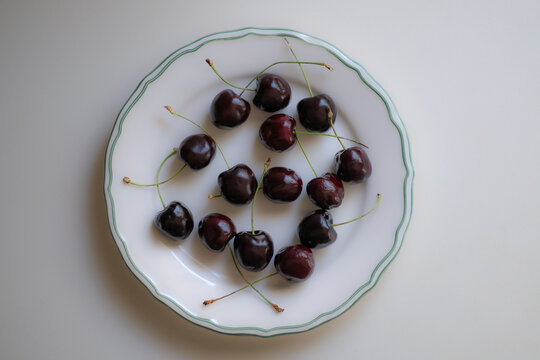 Fresh dark sweet cherries with green stems arranged on a white ceramic plate, simple summer fruit composition on a light background with natural food styling.